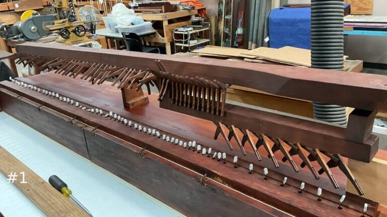 A long, detailed wooden pipe organ windchest component displaying a rows of small metal levers, pins, and valve action mechanics inside a workshop.