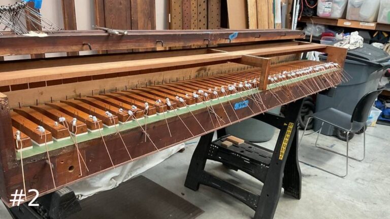A long wooden pipe organ windchest mechanism sits on a black work stand, displaying complex internal rows of wooden blocks, tension springs, and strings.