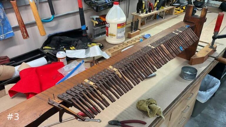 A long wooden organ windchest mechanical assembly featuring rows of closely aligned wooden levers sits clamped on a workshop table.