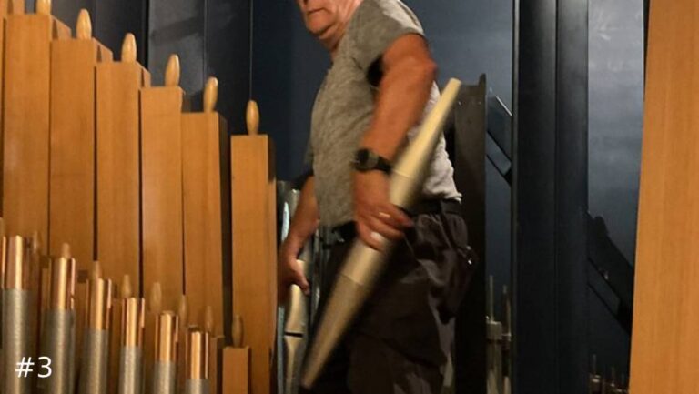 An organ technician carefully guides a long, tapered metal organ pipe downward into place inside a compact organ chamber next to wooden pipes.