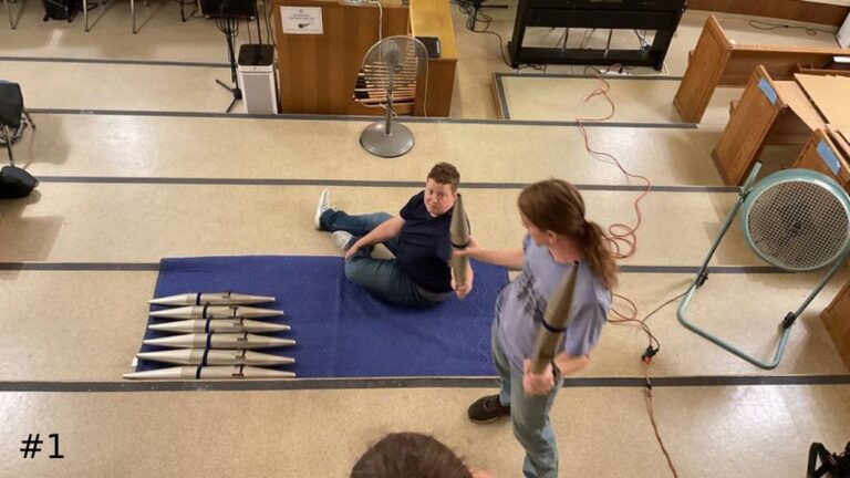 High-angle view of two organ technicians in a church room handing down a zinc organ pipe, next to a blue mat holding a graduated row of metal pipes.