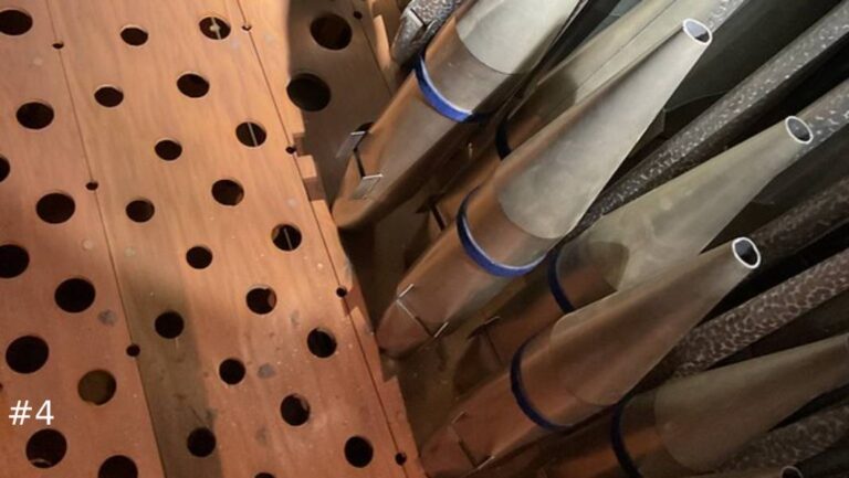 High-angle close-up view of tapered metal organ pipes fitted into place alongside a wooden toeboard rack with empty circular mounting holes.