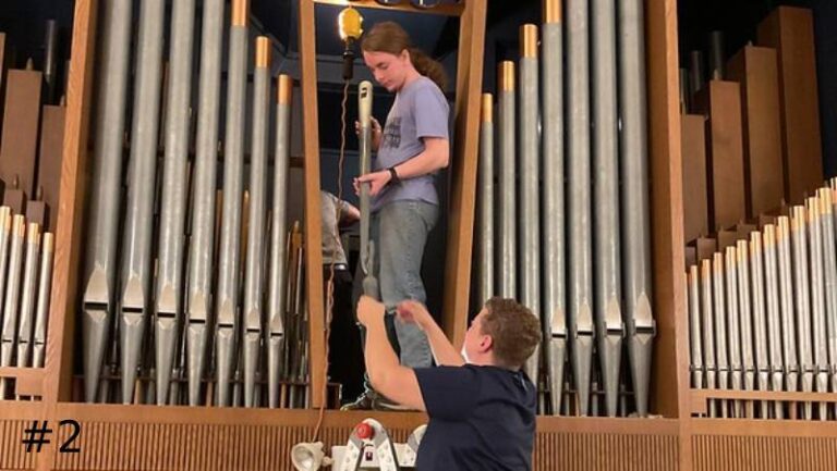 Two organ technicians carefully guide a tall zinc facade pipe up into place within a large pipe organ structural casework framework.
