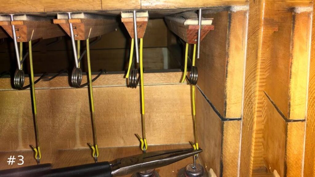 Close-up view inside a wooden organ console casing showing a row of heavy metal coil return springs connected to vertical rods, with needle-nosed pliers resting below