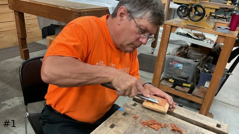 An organ technician sits at a workbench using a chisel to carve a small block of wood, surrounded by wood shavings