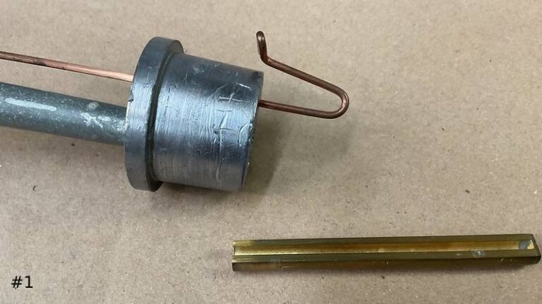 Close-up of a disassembled metal organ reed pipe mechanism on a work surface, featuring a lead block with a copper tuning wire alongside a separate brass shallot channel.