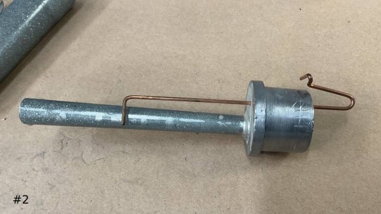Close-up of a partially assembled metal organ reed pipe component on a workbench, showing the copper tuning wire running alongside the long lead tube section.