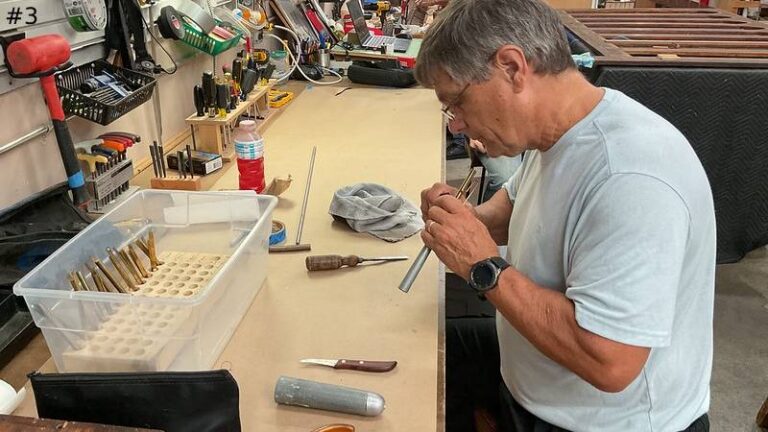 An organ technician blows into a small metal reed pipe component at a workbench filled with tools