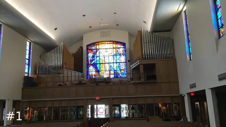 Wide view of a church sanctuary from the main floor looking up at an elevated rear balcony balcony containing a split pipe organ structure