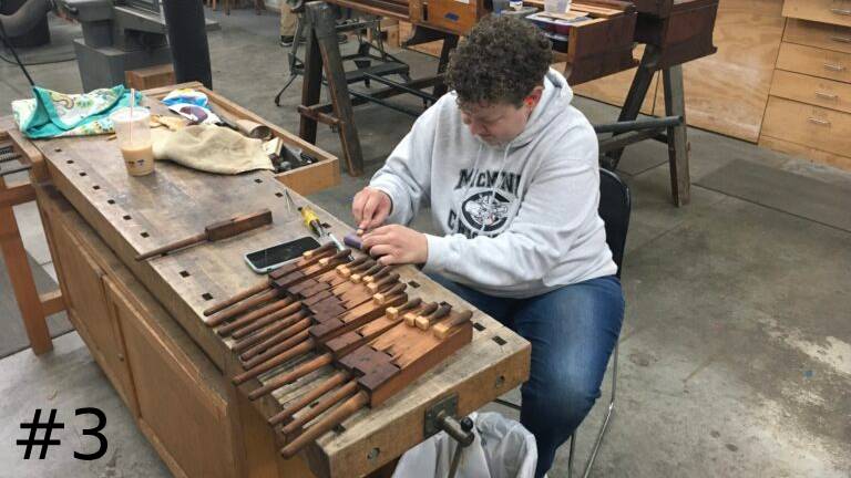 An organ technician sits at a workbench, carefully adjusting a row of small wooden pipe components.