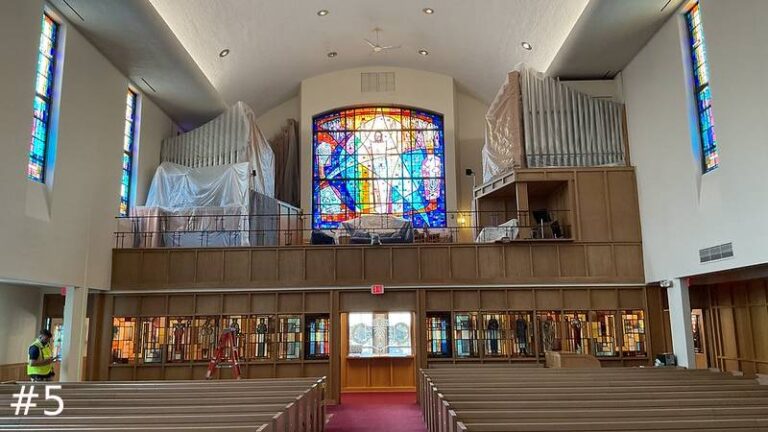 Wide view of a church sanctuary looking toward a rear balcony gallery where the left and right pipe organ structural sections are covered in clear protective plastic sheeting