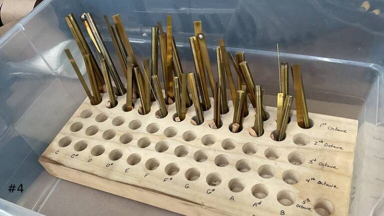 Close-up of numerous brass organ pipe shallots arranged in a wooden block grid labeled by note names and octaves inside a clear plastic container.
