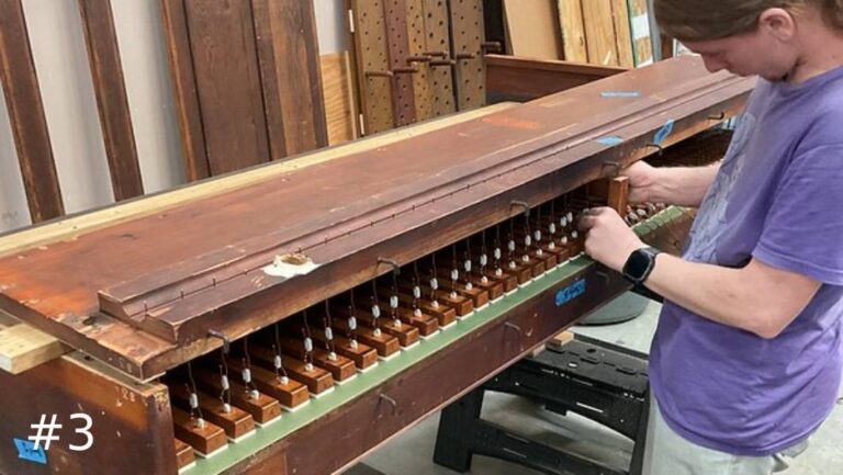 An organ technician inspects the long row of small wooden valve components inside a large pipe organ windchest frame.