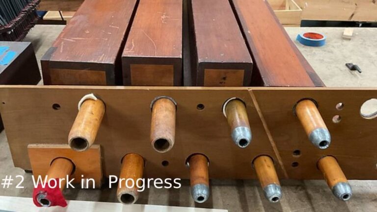 A row of square wooden organ pipes rests in a wooden rack with round protruding pipe feet
