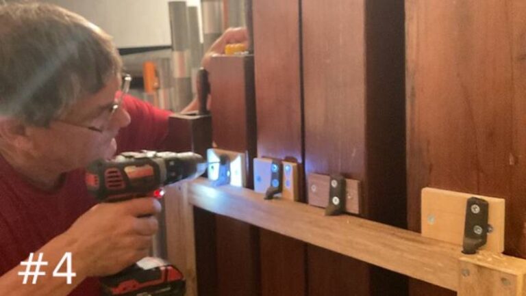 An organ technician holds a cordless drill to drive a screw into a wooden support rail running across several large, vertical wooden organ pipes