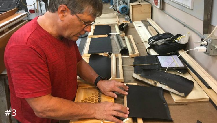 A Leek Pipe Organ Company technician in a red shirt, face mask, and work gloves uses a sanding block to smooth a wooden component on a workshop table.